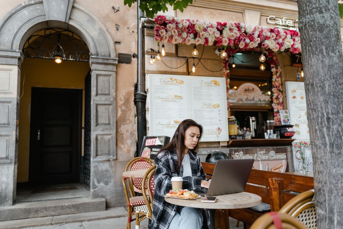 Eine Frau arbeitet mit Laptop und Kaffee in einem Straßencafé voller Blumen.