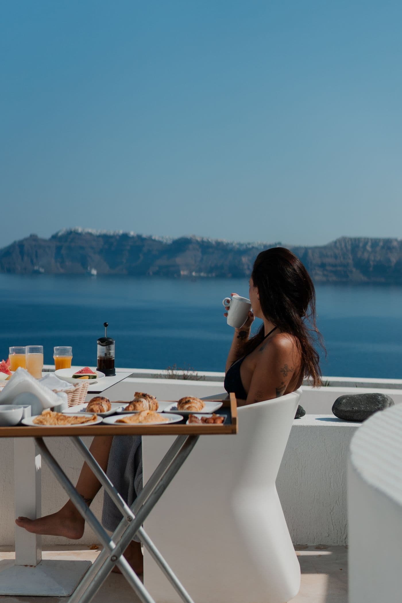 Ein Frühstückstisch mit Kaffee auf einer Terrasse am Meer, eine Frau schaut aufs Wasser.